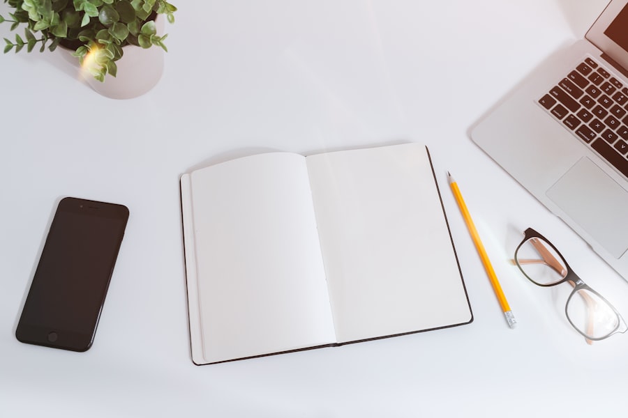 Open notebook on a white desk with a smartphone, pencil, glasses, plant, and laptop.